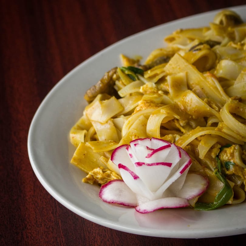 Curry noodles with chicken and vegetables, garnished with a carved radish flower on a white plate.