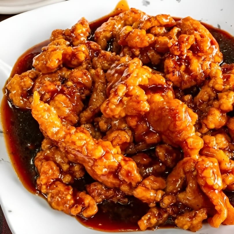 Close-up of crispy orange chicken pieces coated in a rich, dark sauce on a white plate.