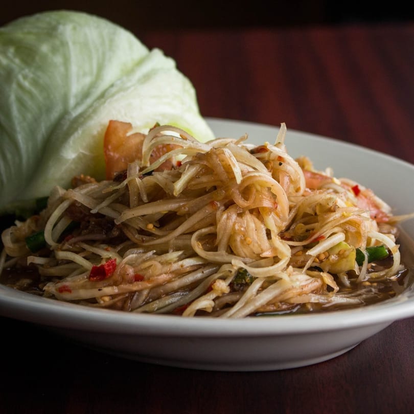 Close-up of a papaya salad with chili flakes, tomatoes, and a side of lettuce.